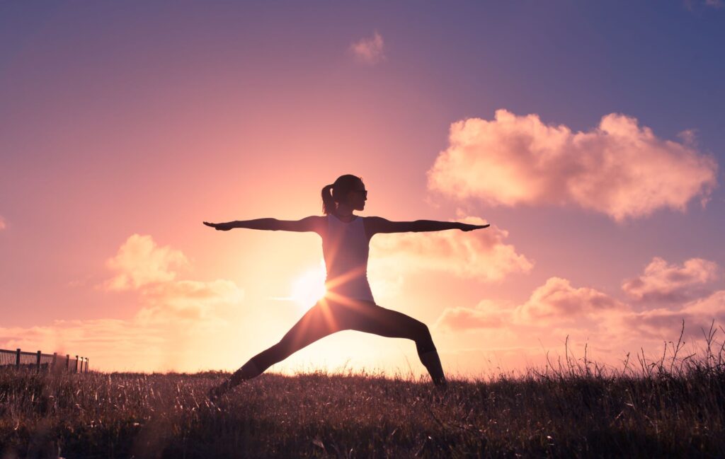 Frau praktiziert Yoga in der Kriegerhaltung vor einem atemberaubenden Sonnenuntergang, symbolisiert Harmonie und innere Ruhe.