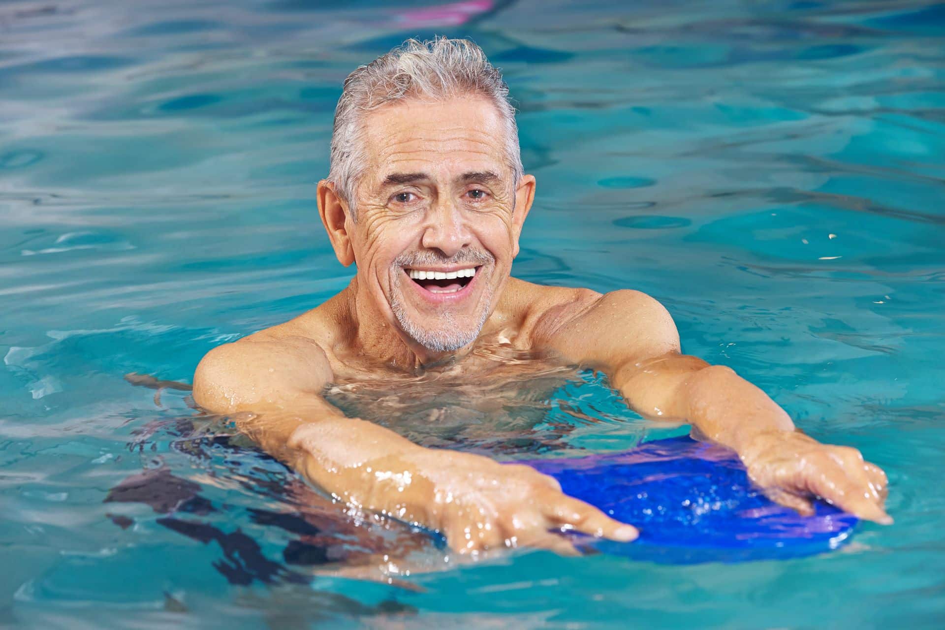 Älterer Mann lächelt und hält ein Schwimmbrett im Pool.