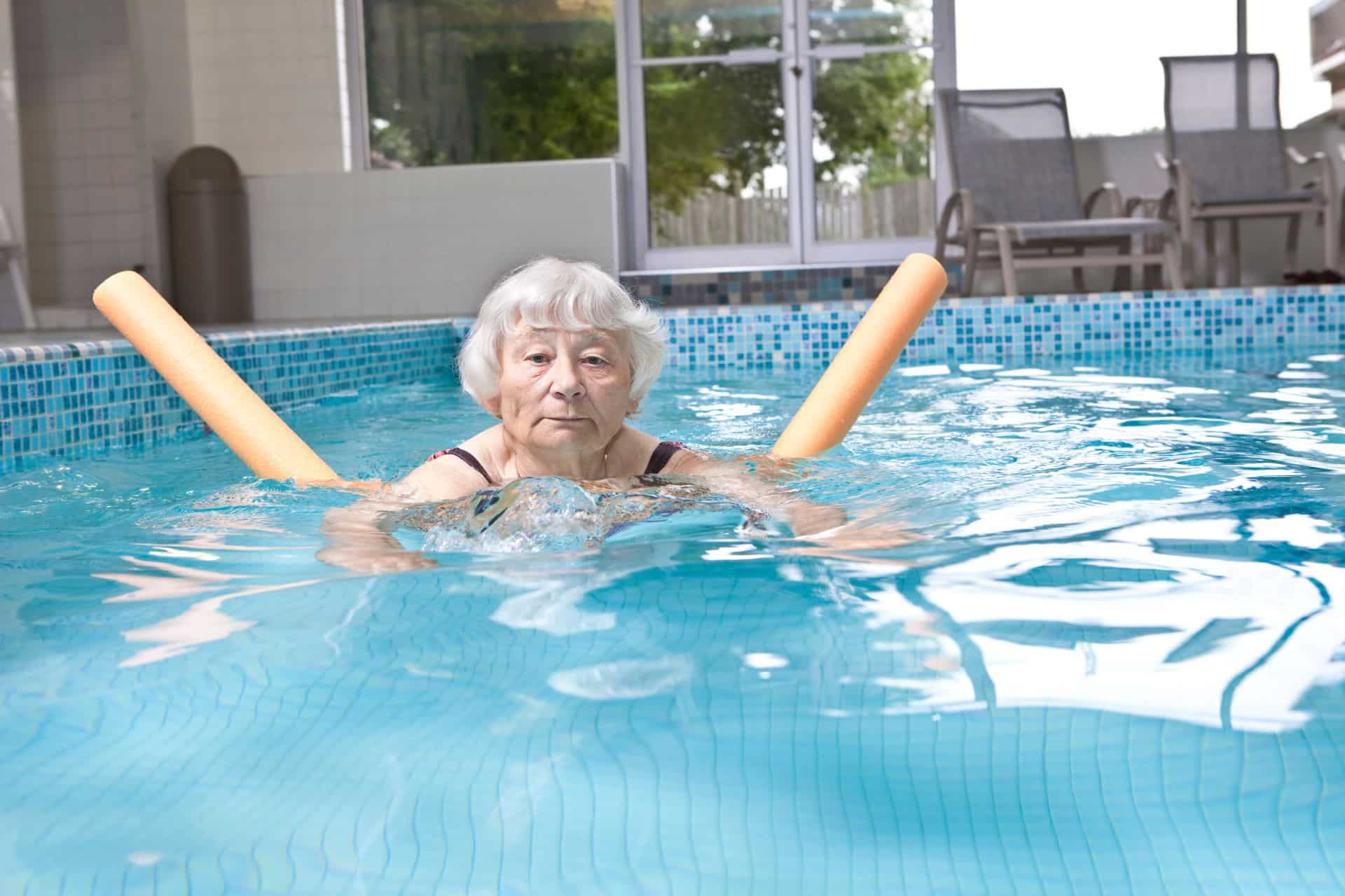 Ältere Dame schwimmt mit einer Poolnudel in einem Innenpool.